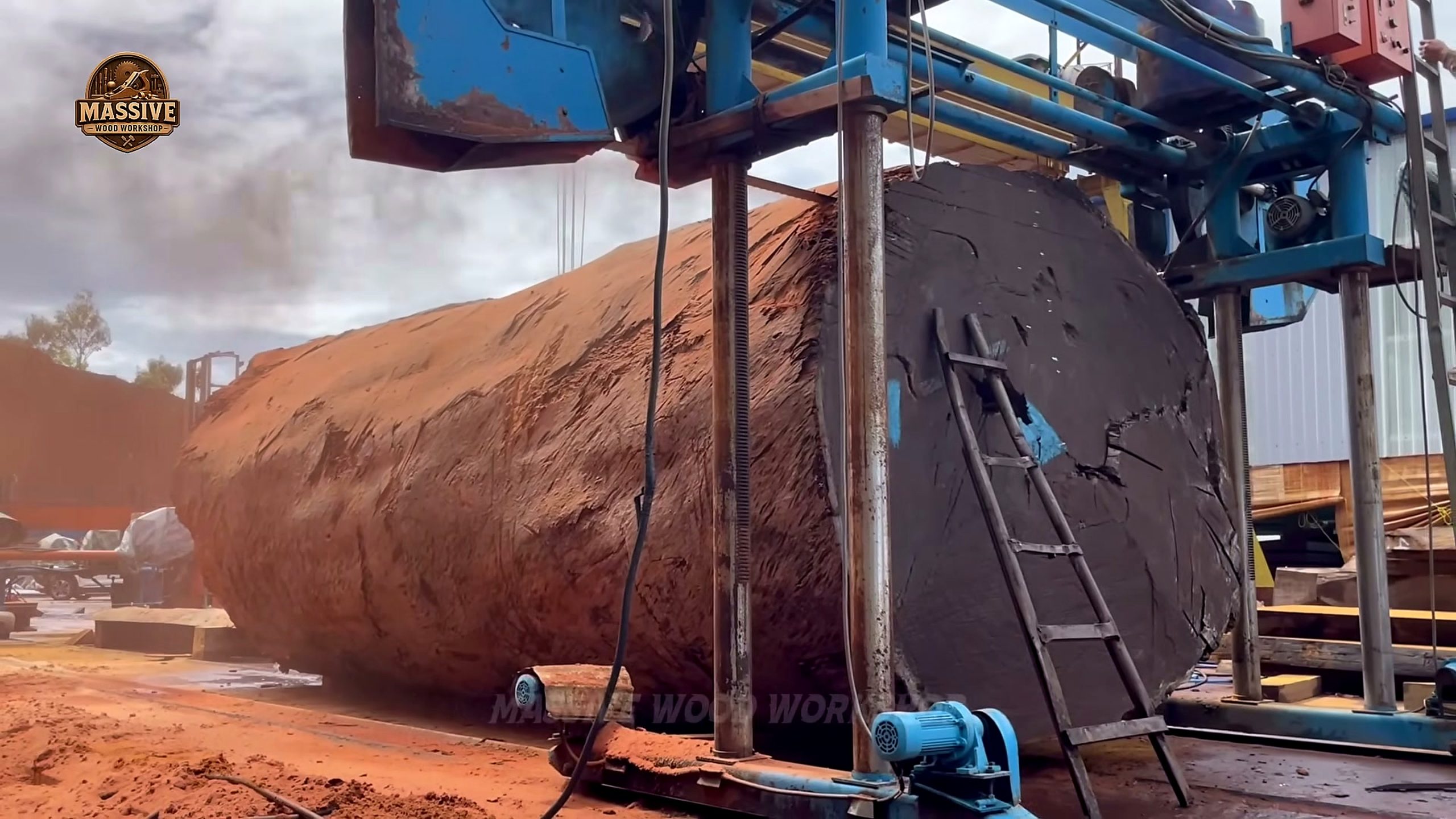 giant timbers under the blue frame of a travelling band mill with a ladder against the uprights